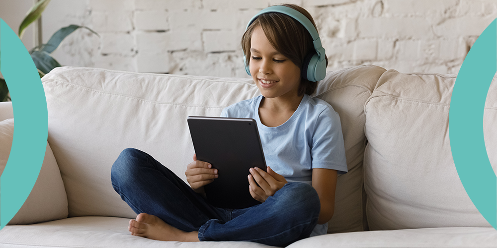Picture of a young child sitting cross legged on a sofa, wearing headphones and reading from a tablet.